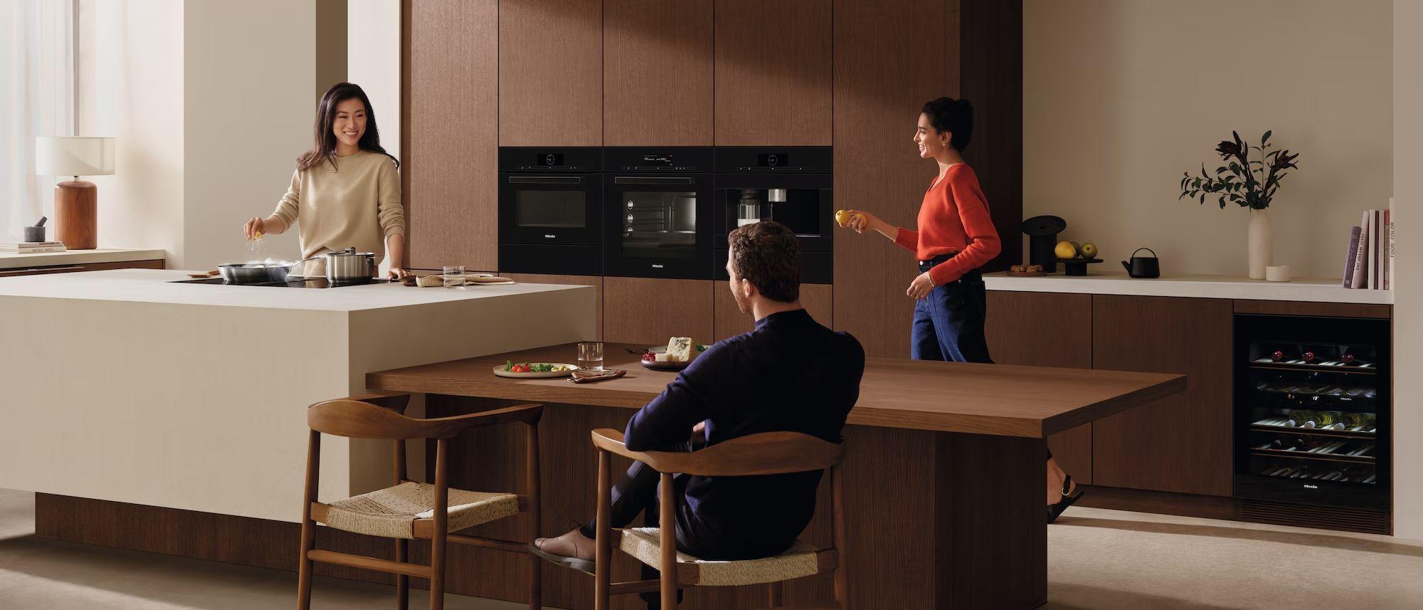 Miele Küchentechnik in der Wohnküche – helle Kochinsel mit Kochgeschirr, warme Holzfronten, Miele Einbaugeräte (Backöfen und Weinschrank) sowie integrierter Esstisch in offenem Raumkonzept