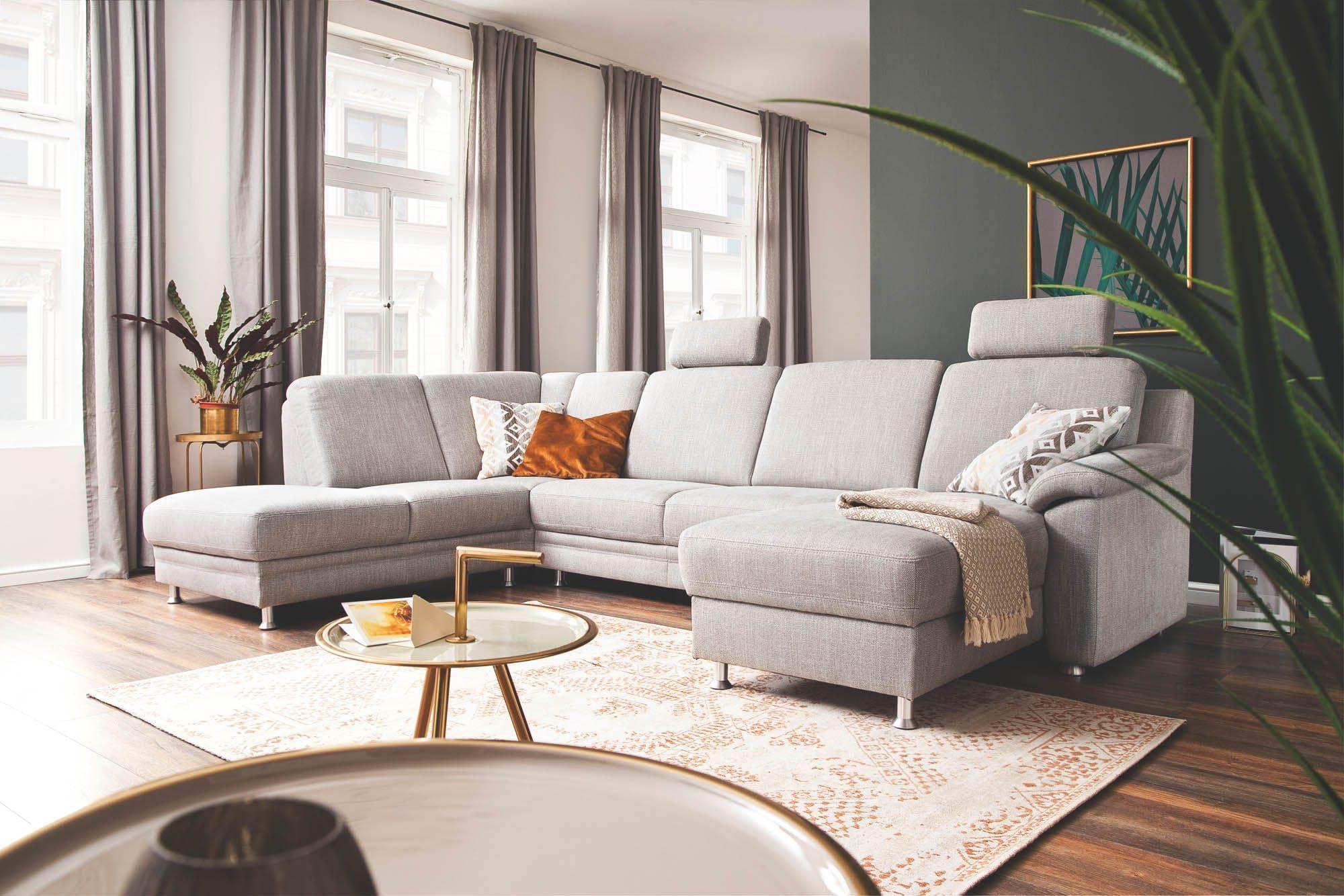 Helles Wohnzimmer mit großer hellgrauer Eckcouch samt Kopfstützen, Kissen und Decke, dazu runder Beistelltisch, gemusterter Teppich, hohe Fenster mit Vorhängen und Pflanzen.