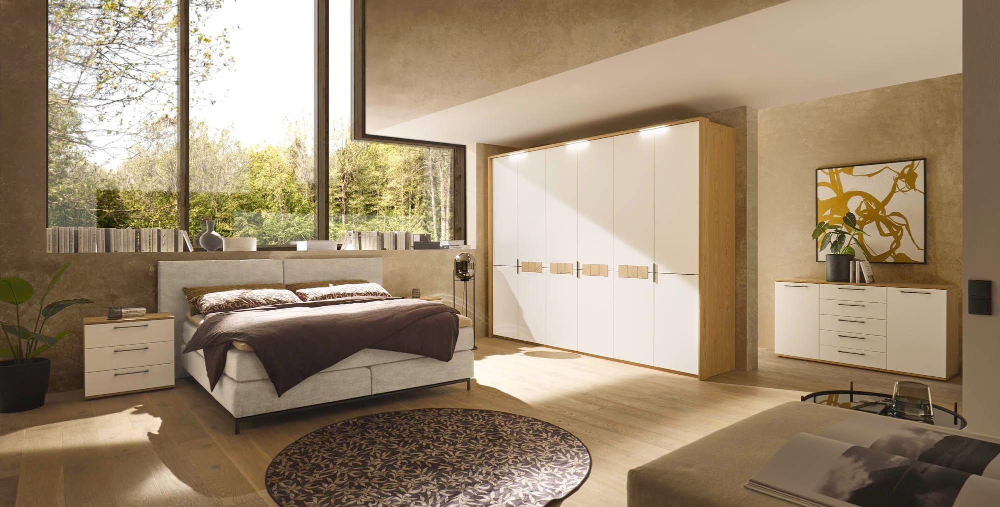 Calvia Schlafzimmer komplett in Weiß und Eiche mit Polsterbett in Grau, Drehtürenschrank mit Holzgriffen, Nachttisch und Sideboard in sonnendurchflutetem Raum mit Waldblick