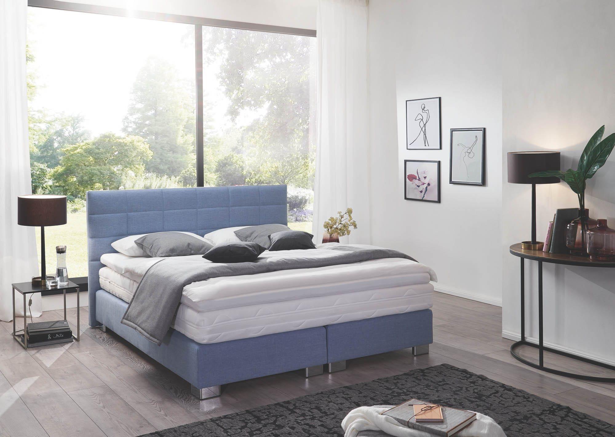 Ambience Boxspringbett in Jeansblau mit quadratisch gestepptem Kopfteil und Chromfüßen, graue Kissen und Tagesdecke in hellem Schlafzimmer mit Gartenblick