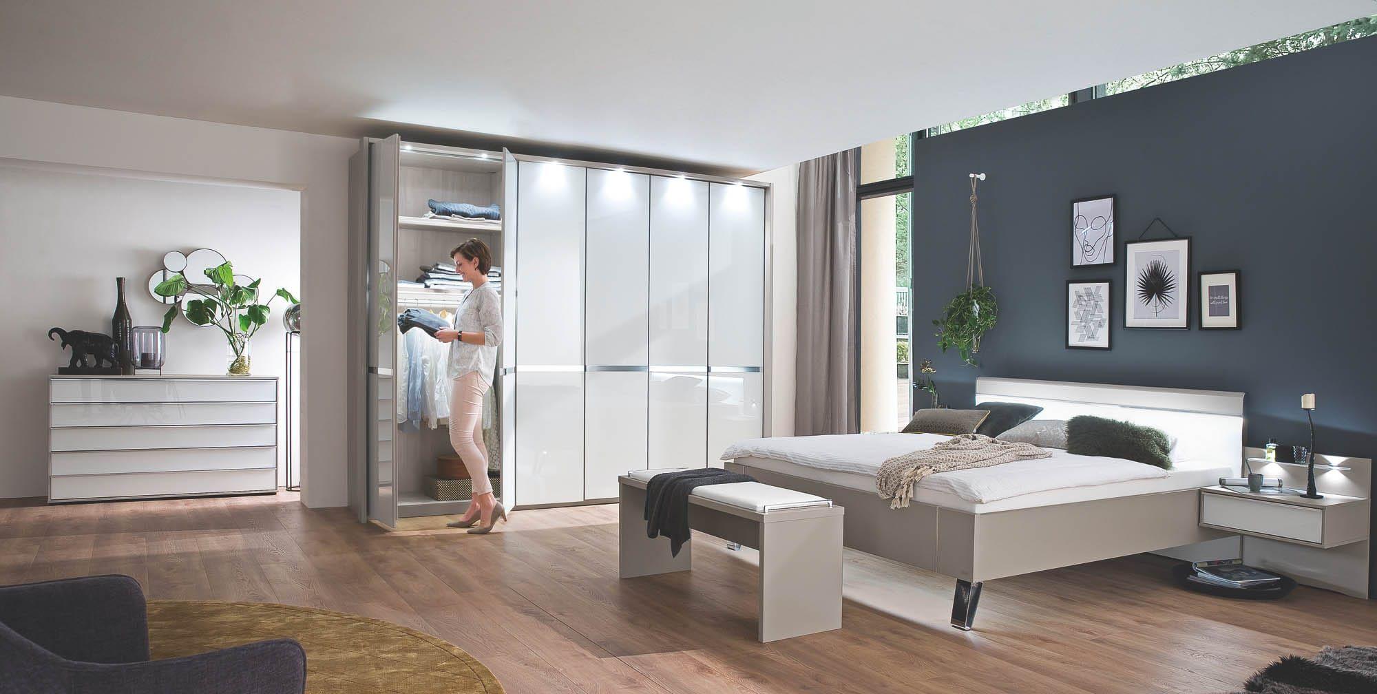 Wiemann Carberry Schlafzimmer in Kieselgrau und Weißglas mit beleuchtetem Falttürenschrank, Bett mit LED-Kopfteil, Kommode und Sitzbank in modernem Ambiente mit petrolblauer Akzentwand
