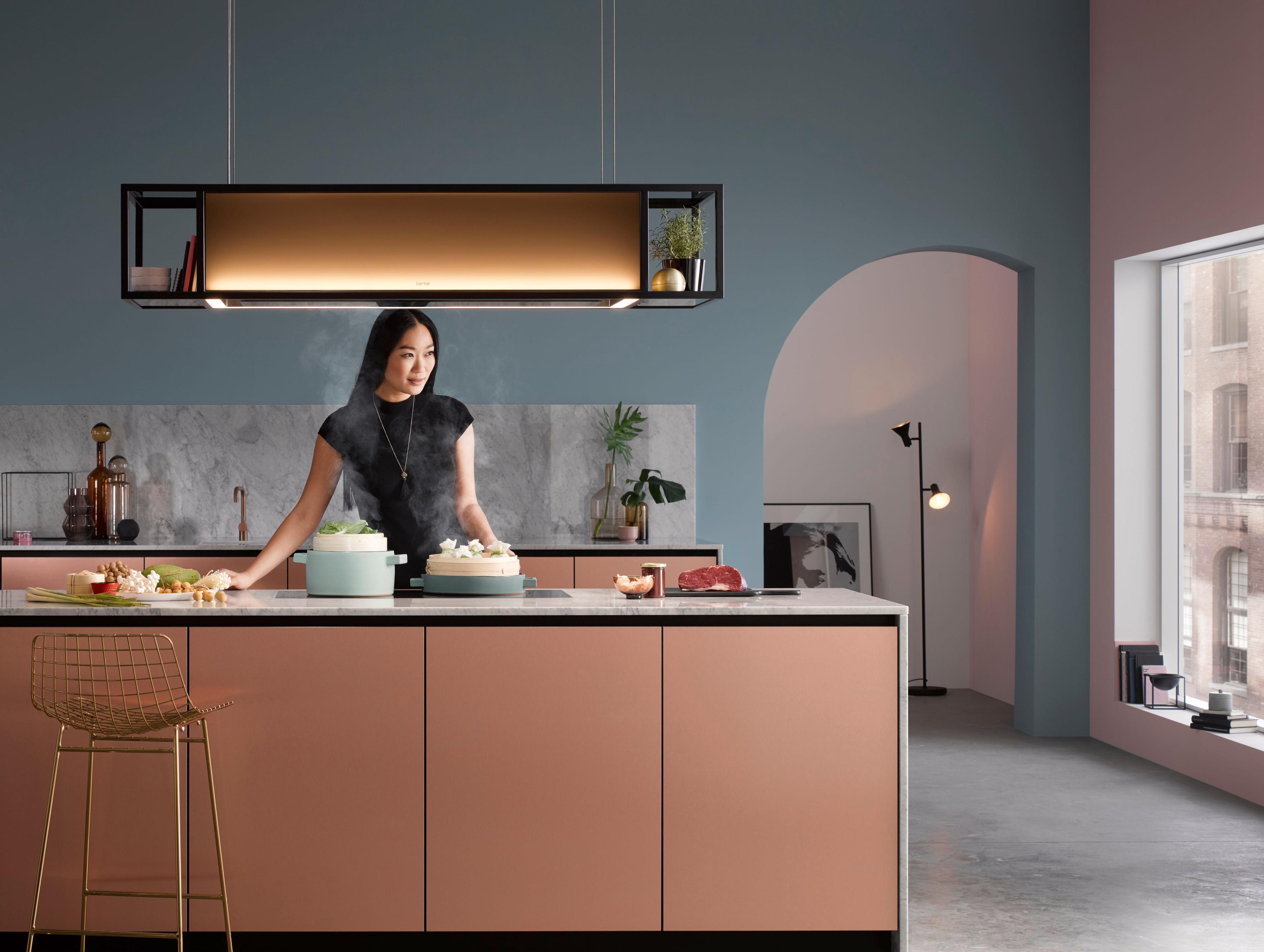 berbel Skyline Frame Dunstabzug über moderner Kücheninsel – offene Rahmenkonstruktion mit integrierter Beleuchtung; Person kocht mit dampfenden Töpfen in farbig gestaltetem Wohnküchenraum