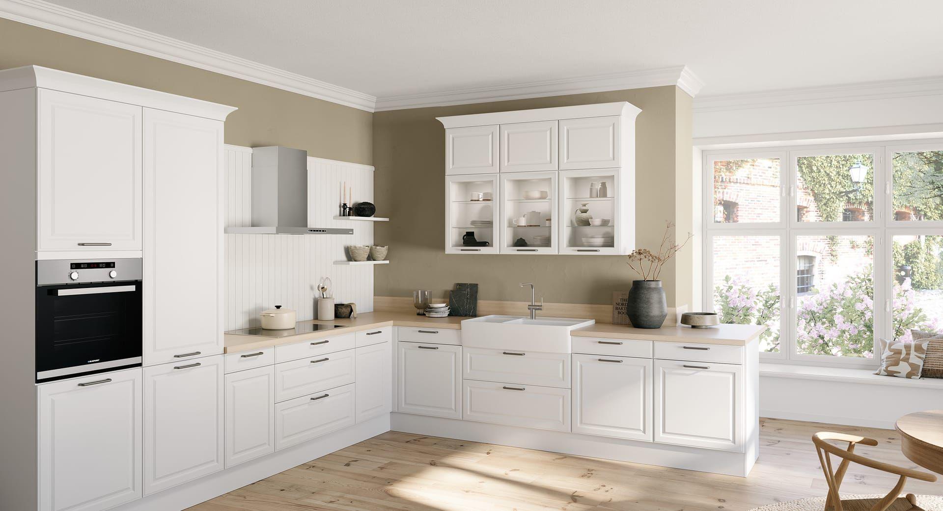 Häcker Küche Toskana in Kristallweiß – klassische Landhausküche in L-Planung mit Rahmenfronten, Vitrinen, Holzarbeitsplatte und großer Keramikspüle in einem hellen Raum mit Fensterbank-Sitzplatz