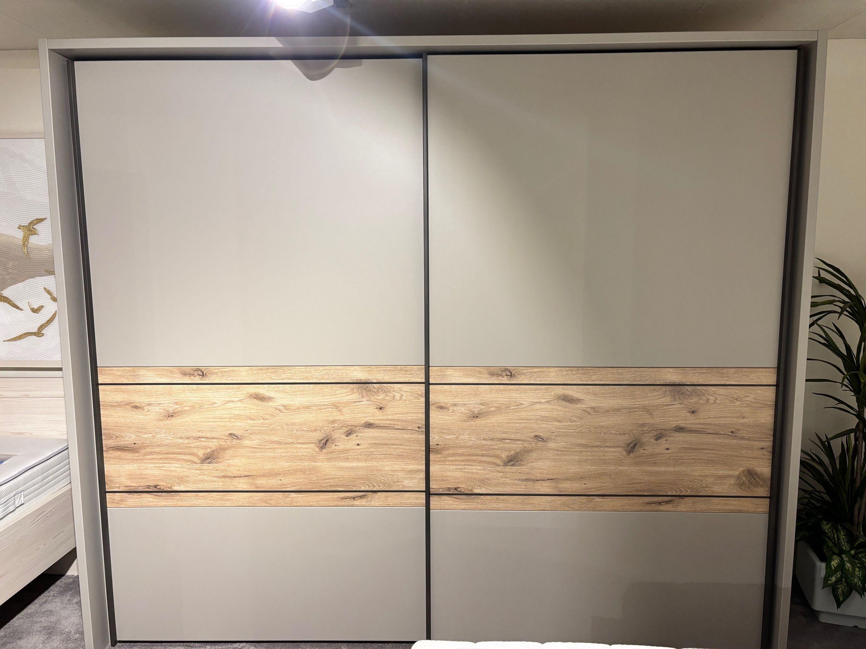Nahaufnahme des Schwebetürenschranks in Grau mit Eiche-Holzdekor-Einsatz, zweitürig mit schwarzen Rahmenleisten.