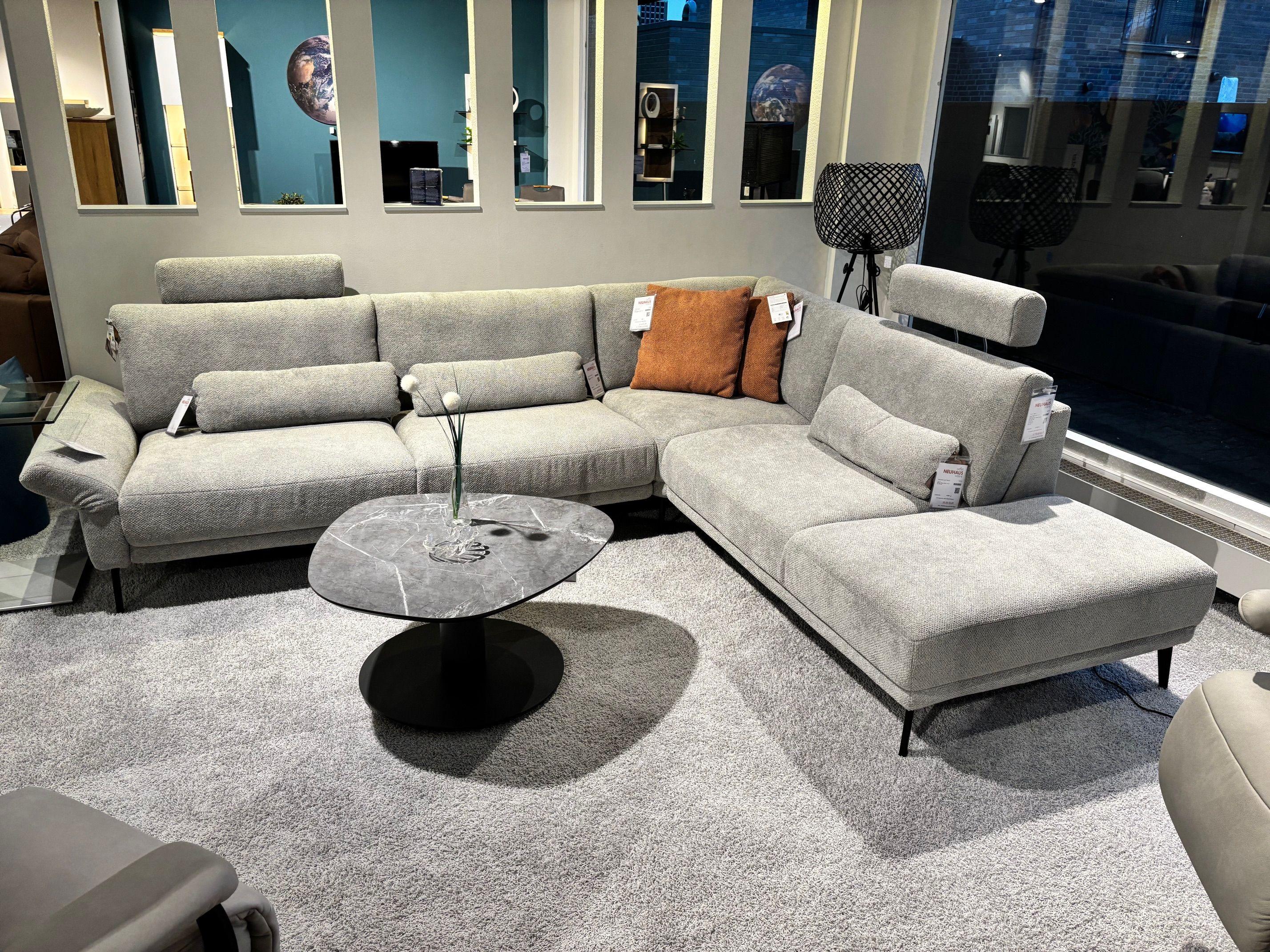 Hellgraue Eckcouch mit verstellbaren Kopfstützen, cognacfarbenen Kissen und rundem Couchtisch in Marmoroptik auf Hochflorteppich.